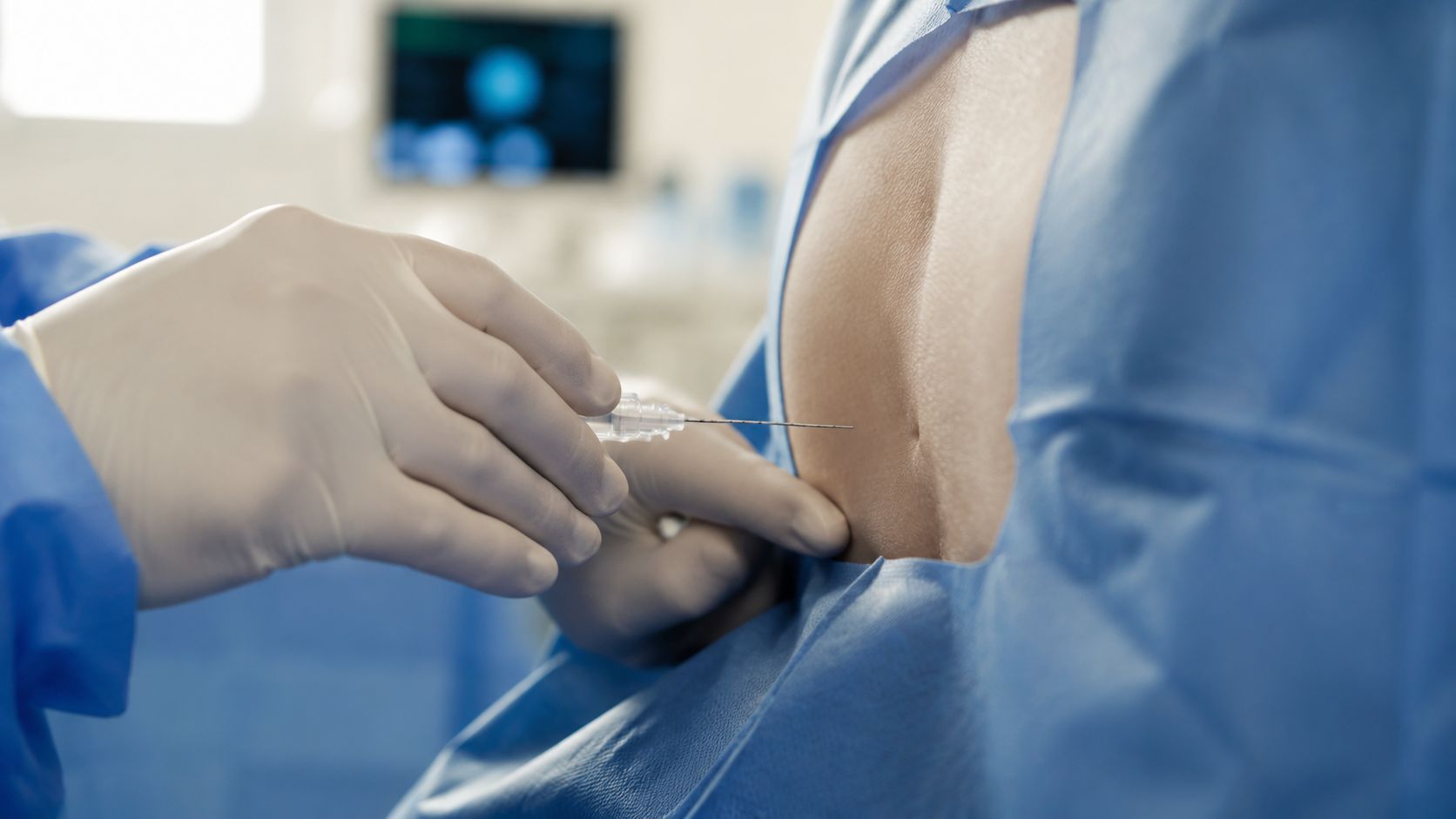 A medical professional in gloves performs a spinal procedure on a patient's back in a clinical setting.