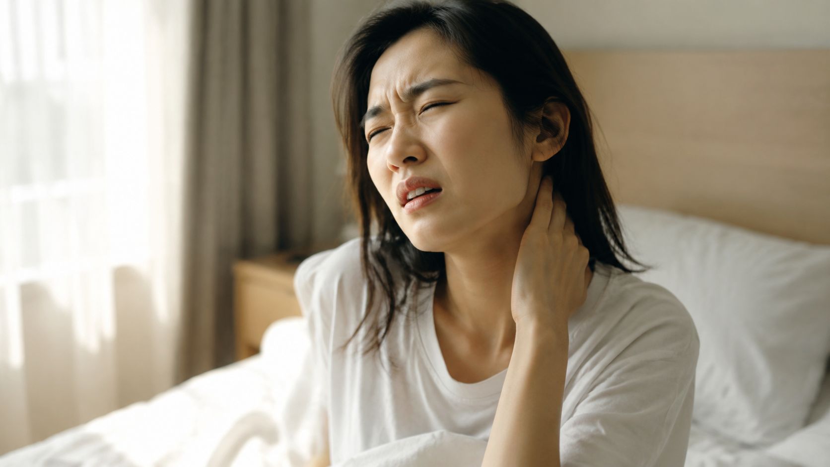 A young Asian woman sitting on a bed in pain while holding her sore neck.